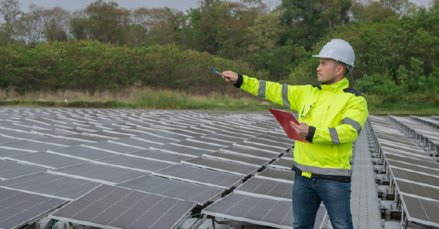 A man wearing a hard hat and yellow jacket stands confidently in front of a row of solar panels, showcasing renewable energy