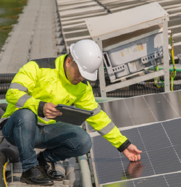 A man in a yellow jacket and hard hat inspects solar panels using a tablet for monitoring and assessment